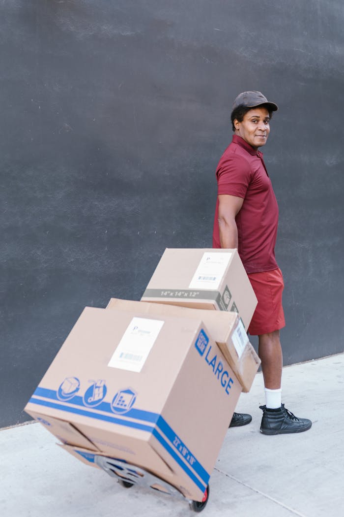 A delivery man in uniform with packages on a hand truck against a wall.